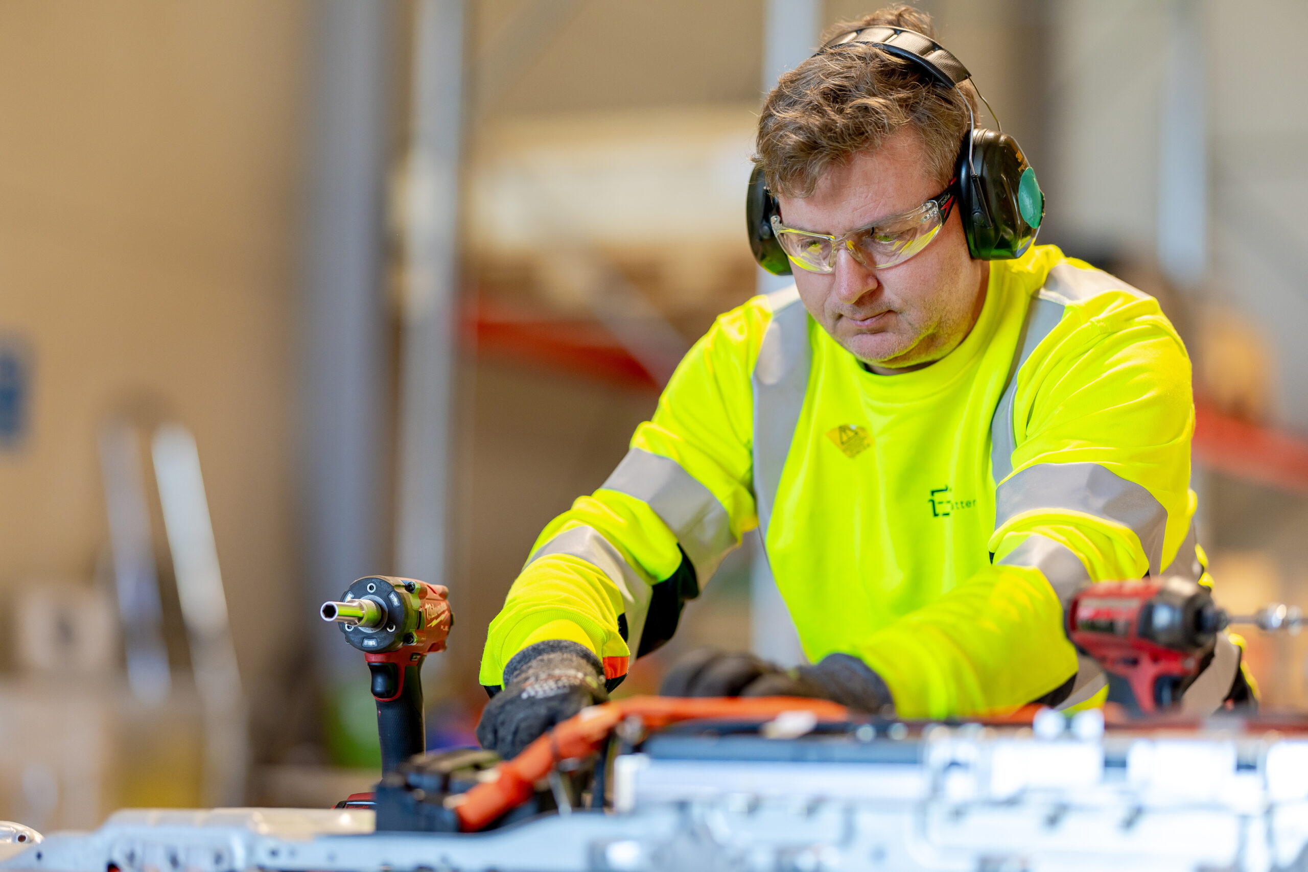 Industriarbeider i gul vernejakke og hørselvern som bruker elektroverktøy i verksted